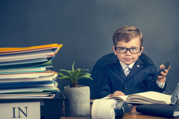 Young boy dressed in a suit working at a large desk. He is using a mobile phone. The desk has a calculator, Filofax, phone and clock. He is very busy and there is a lot of work on his desk. He is wearing glasses and looks like an accountant.
